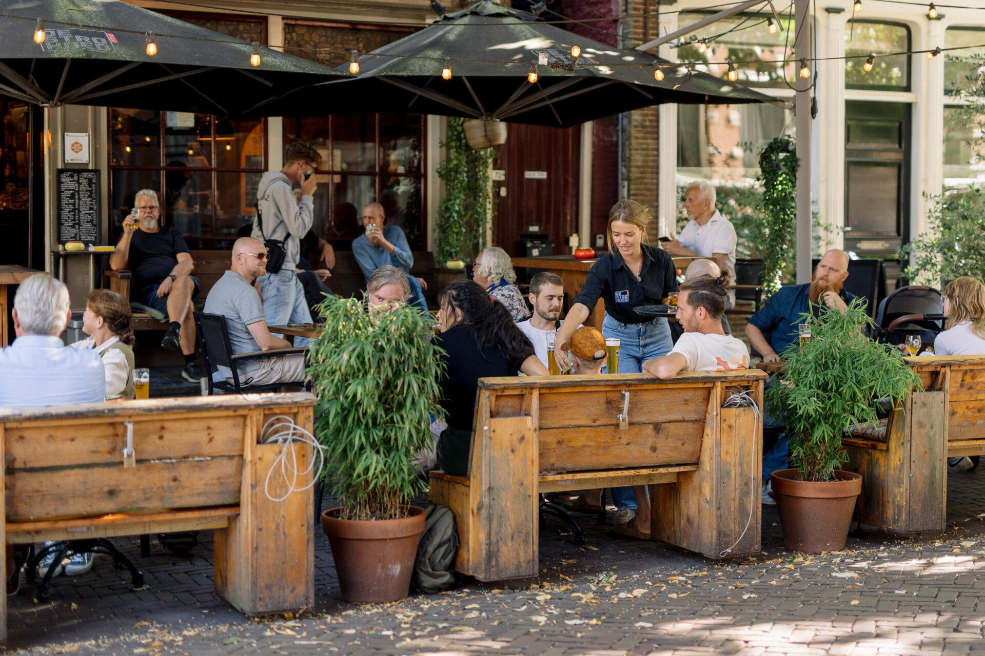 Het koele terras van Café de Strohoed, waar mensen in de schaduw afkoelen op een warme zomerdag.
