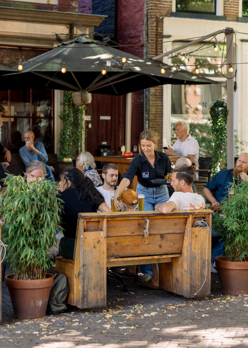 Het koele terras van Café de Strohoed, waar mensen in de schaduw afkoelen op een warme zomerdag.