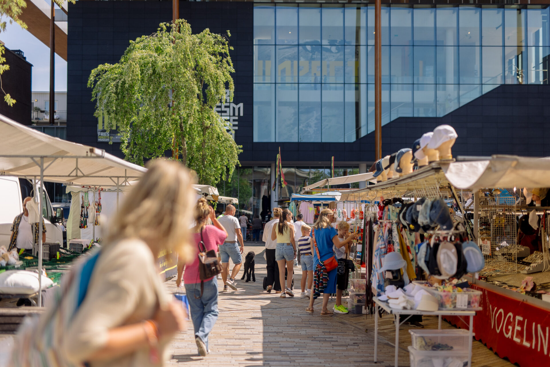 Winkelende mensen op de vrijdagmarkt op het Wilhelminaplein met op de achtergrond het Fries Museum.