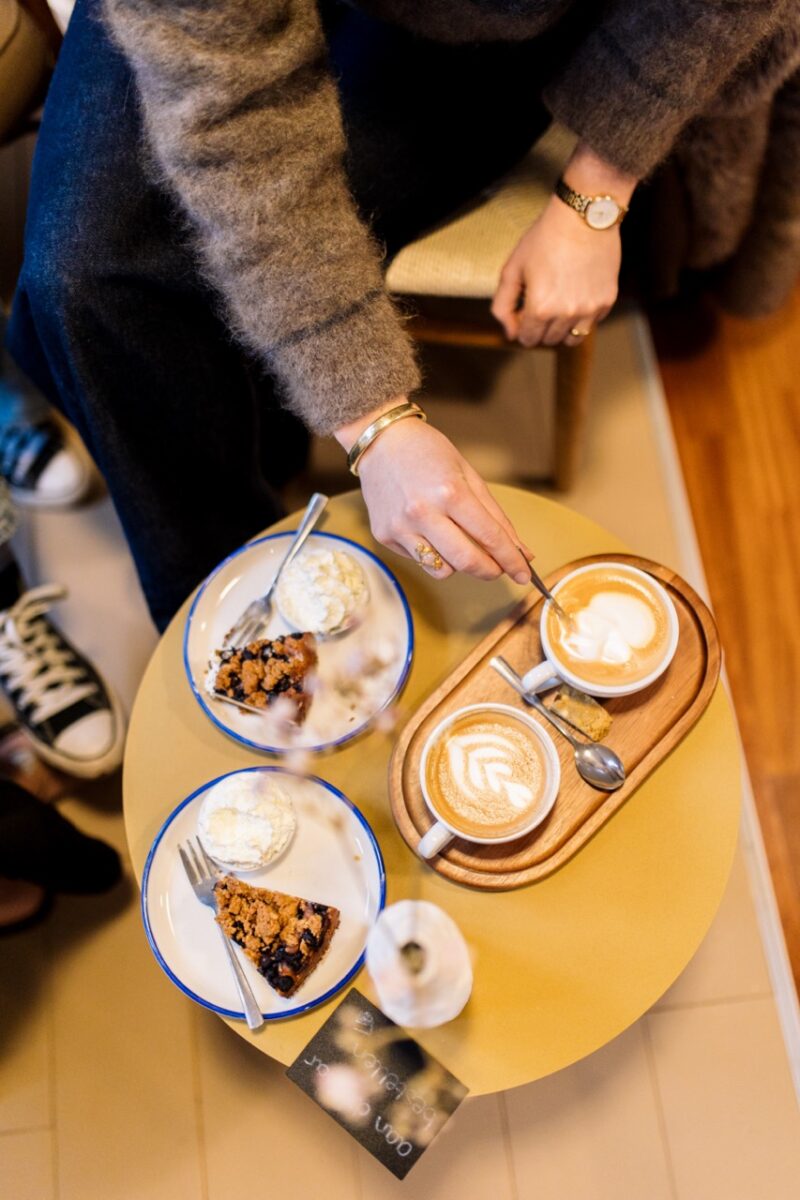 Er wordt in een cappuccino geroerd en er staat taart op tafel