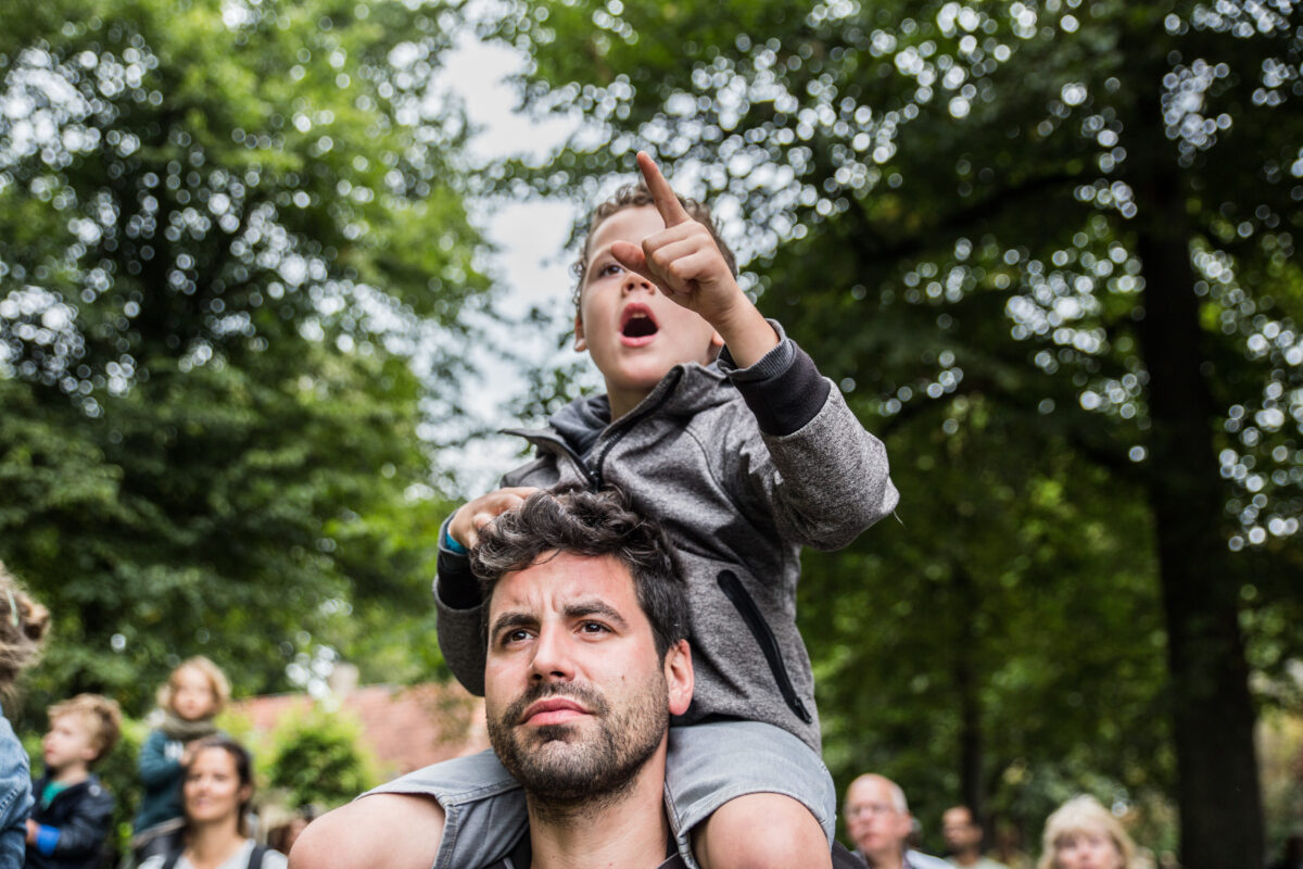 Jongetje zit op de schouders van zijn vader en wijst naar iets wat hij ziet.