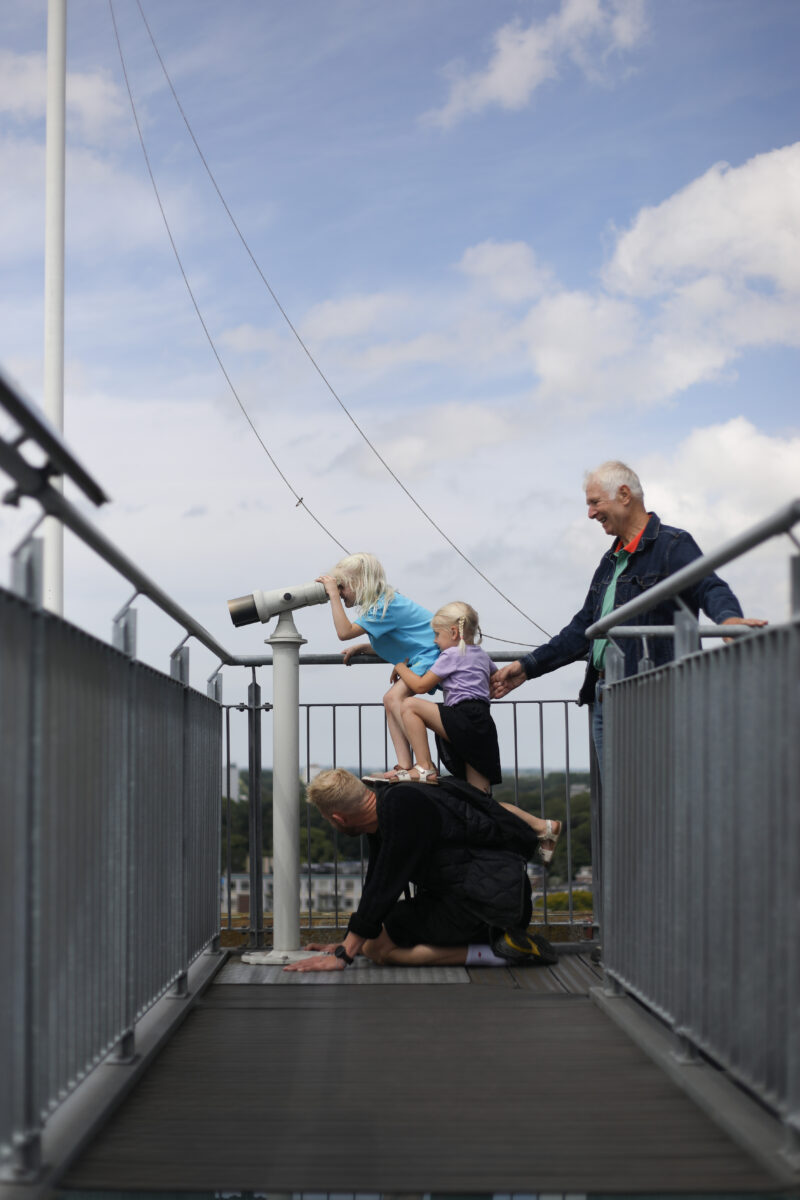 Opa, vader en twee dochters proberen door de telescoop op de Oldehove te kijken. De dochters staan op de rug van hun vader, terwijl die op handen en knieën op de vloer zit.