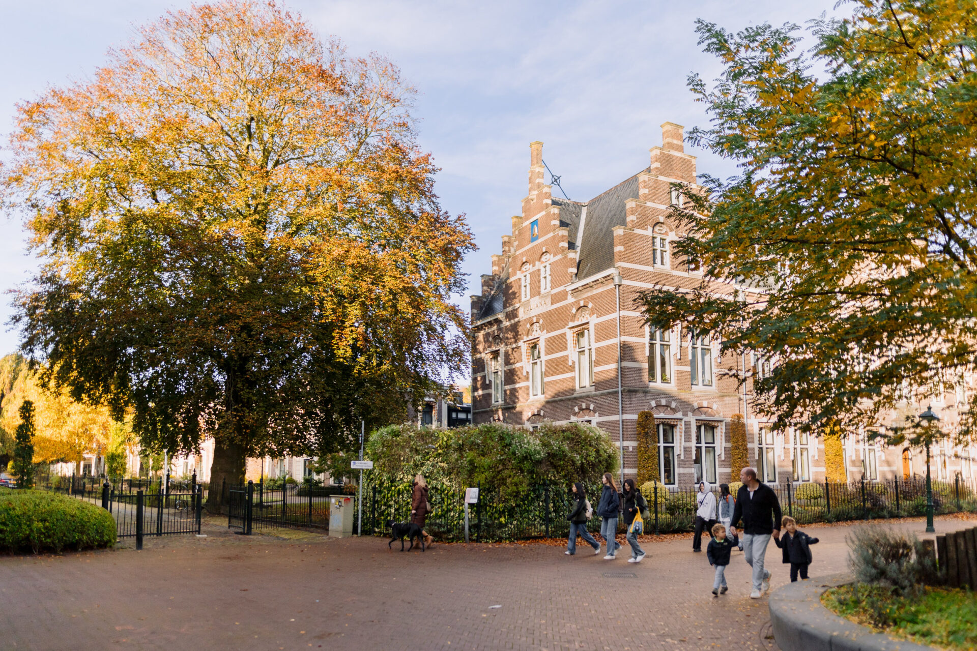 Ouders met kinderen wandelen hand in hand langs het Sint Anthony Gasthuis op een zonnige herfstdag. De bomen zijn oranje bruin gekleurd.